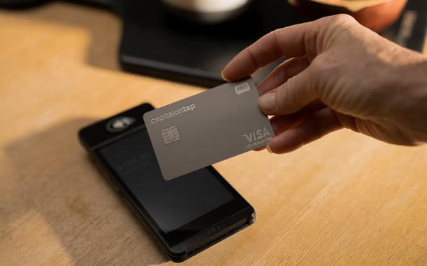Hand Tapping A Capital On Tap Pro Metal Visa Card Against A Contactless Card Reader On A Wooden Counter