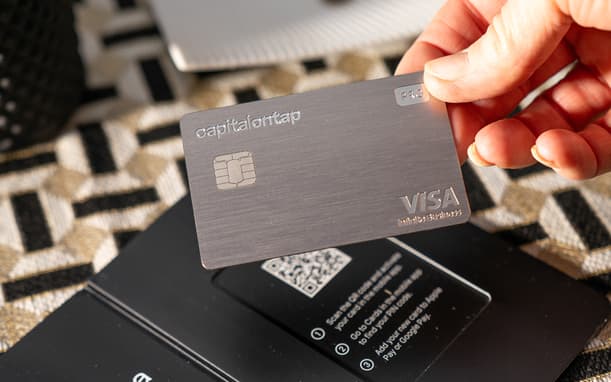 A Hand Holding The Pro Metal Card Over A Patterened Table Cloth In What Looks Like A Hotel Room, With A Duish With A Hotel Key In The Background