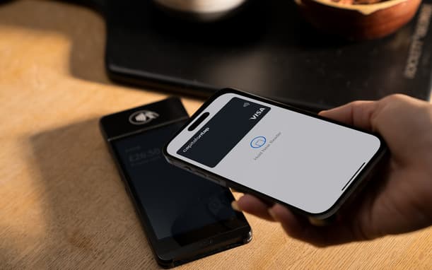 Hand Holding A Phone With Apple Pay Showing The Capital On Tap Visa Card Ready To Tap, Next To A Card Reader Displaying A £26.50 Total