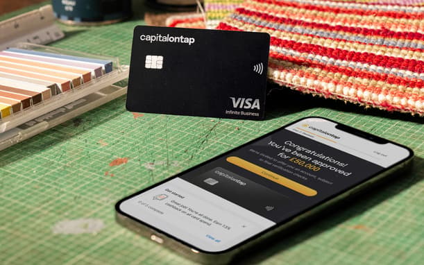 Capital On Tap Visa Card And Phone Showing A £50,000 Credit Approval On The App, Resting On A Designer's Workbench With Fabric Swatches And Paint Tins