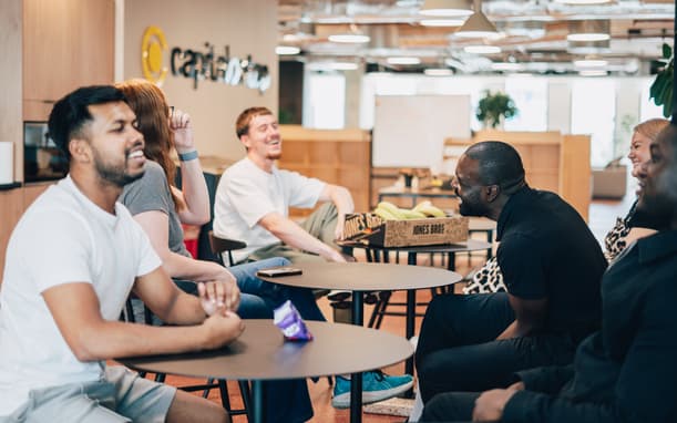 Capital On Tap Colleagues Laughing And Chatting Together Over Snacks In The Office Breakout Area, With The Company Logo On The Wall Behind