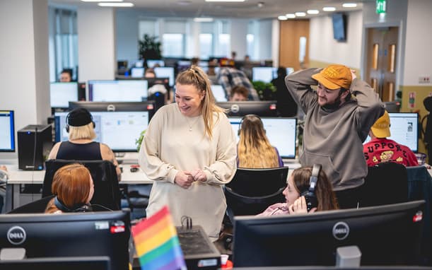 Capital On Tap Employees Smiling And Chatting In A Busy Open Plan Office, With A Small Pride Flag On A Desk In The Foreground