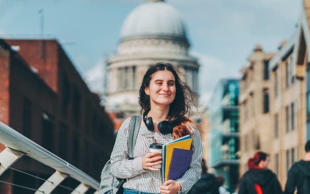 A young woman holding a coffee and books walking and smiling