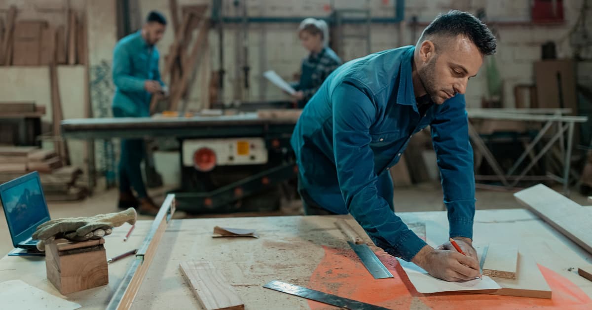 A Man In A Deimn Shirt Writing On A Piece Of Paper, Bending Over A Table Covered In Wood Shavings With A Pair Of Gloves Next To Him