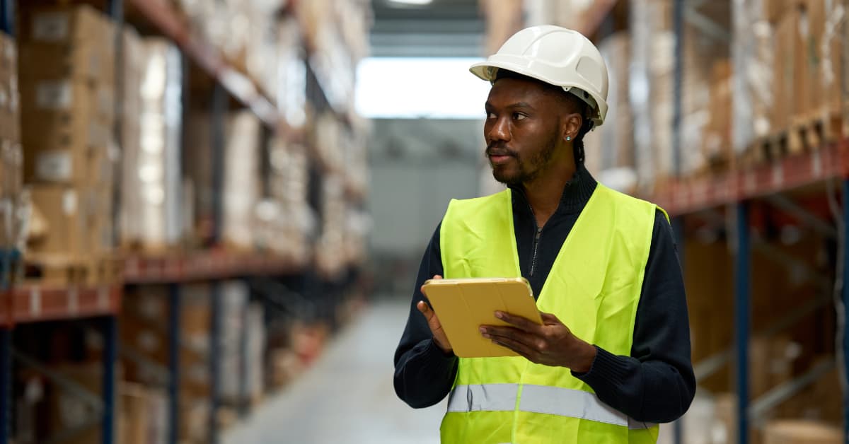 A Man Who Appears To Be In His 30S Stands In A Warehouse In A Hard Hat And A High Vis Vest, Looking At Shelves Filed With Boxes, While Holding A Tablet In His Other Hand.