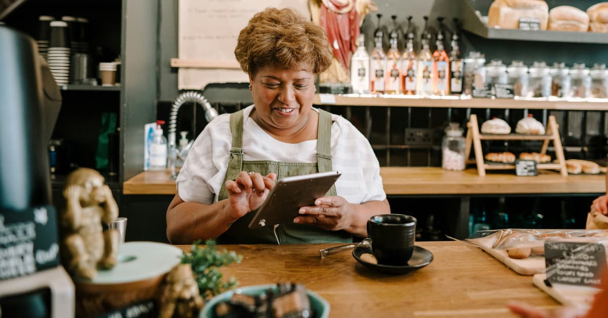 A Small Business Owner In An Apron Stands Behind The Bar In A Cafe Smiling Down At Her Tablet