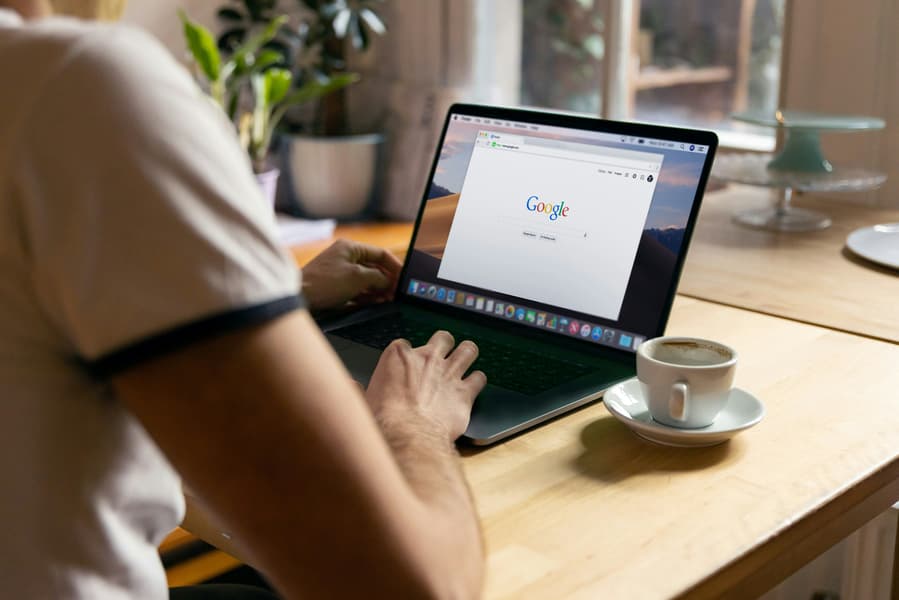 Person Typing On A Laptop With The Google Homepage Open, Next To A Cup Of Coffee On A Wooden Desk