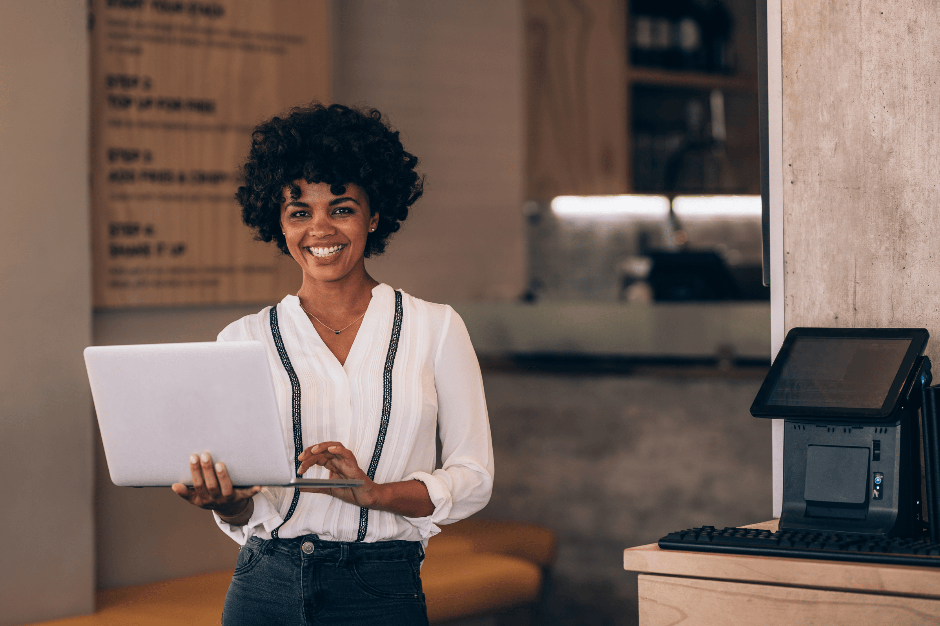 Small Business Owner Standing In A Cafe Near The Till Holding A Laptop And Smiling