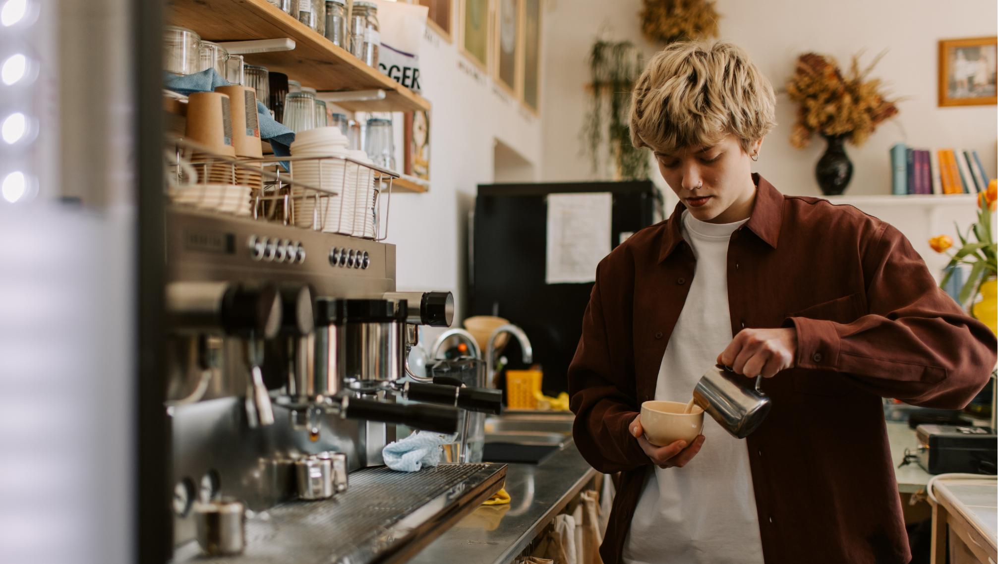 A Barista Wearing A Brown Shirt, Pouring Milk Into A Cappuccino Mug, Standing Beside A Coffee Machine In A Cafe