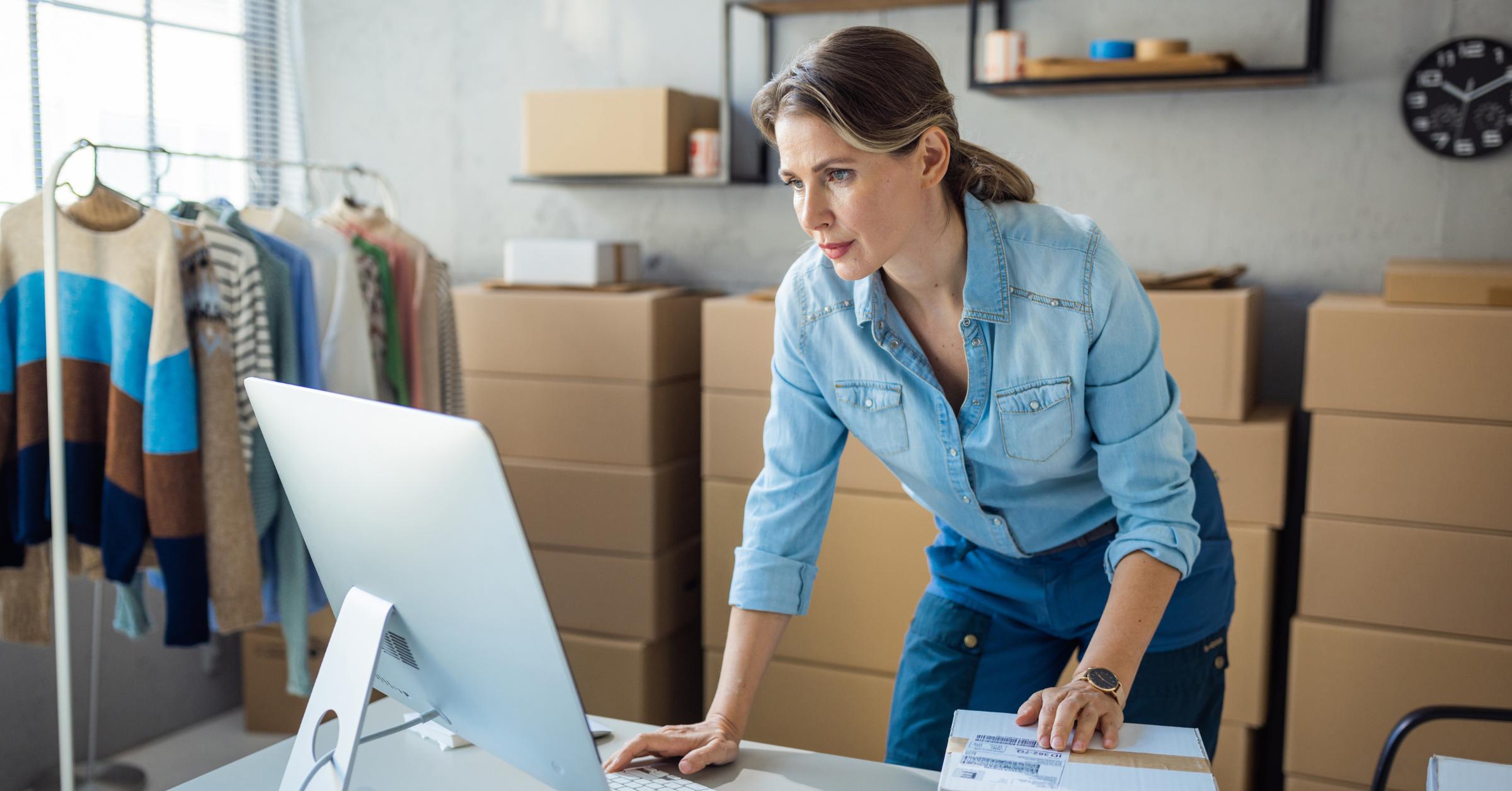 Female Small Business Owner Leans Over A Desk Looking At Her Desktop, In A Room Filled With Boxes And Clothing Rails