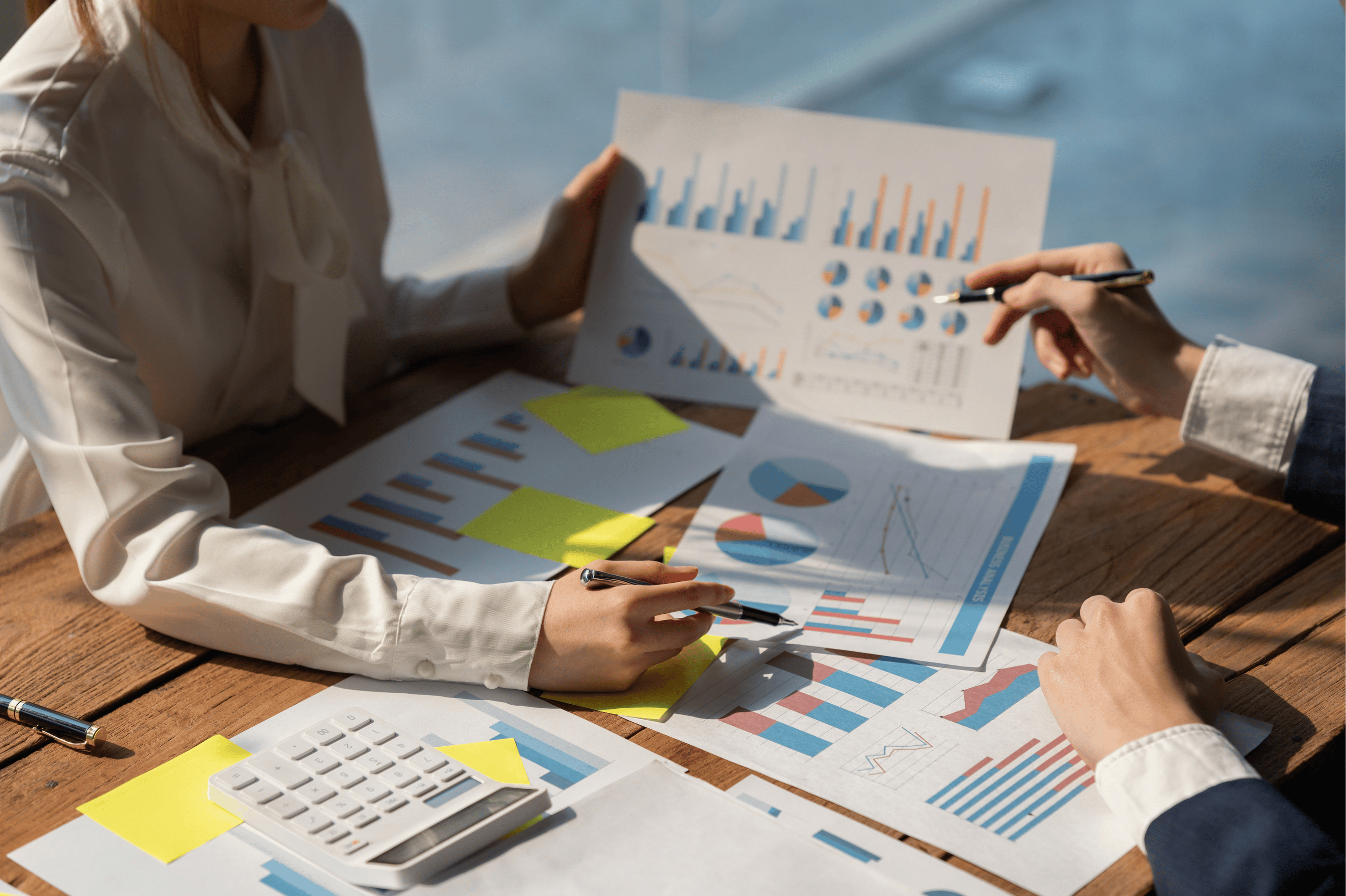 Two Employees Sitting At A Table Covered With Papers Showing Charts And Data, Trying To Figure Out Profit