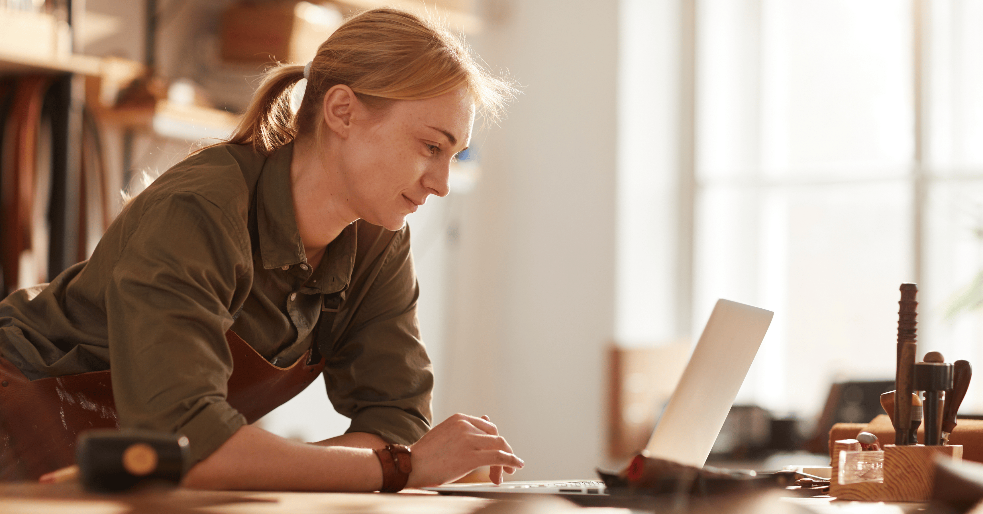 A Small Business Owner Wearing An Apron In Her Woodwork Shop, Leaning Over A Counter Looking At Her Laptop With A Concentrated Look On Her Face
