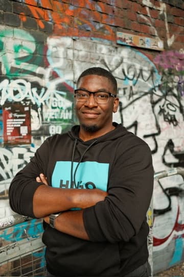 Kyle, Founder Of Social Nerd, Wearing Glasses And A Black Hoodie. He's Smiling, Posing In Front Of A Wall Covered In Graffiti.