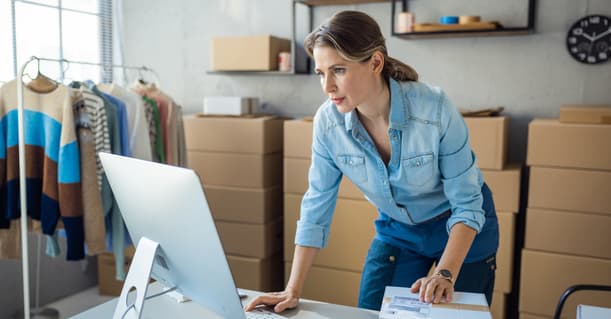Female Small Business Owner Leans Over A Desk Looking At Her Desktop, In A Room Filled With Boxes And Clothing Rails