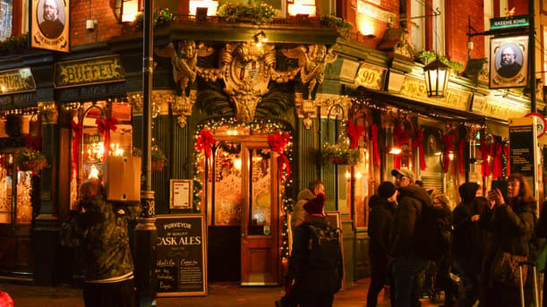 The Salisbury, A Historic London Pub Decorated With Red Bows And Fairy Lights, With Shoppers And A Man Carrying A Box Passing By At Night