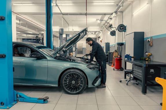 Mechanic In Dark Overalls Working Under The Bonnet Of A Car In A Bright Garage Workshop