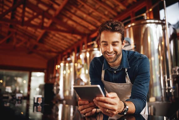 A Man In An Apron Leans Over A Counter In A Brewhouse, Smiling At His Tablet.