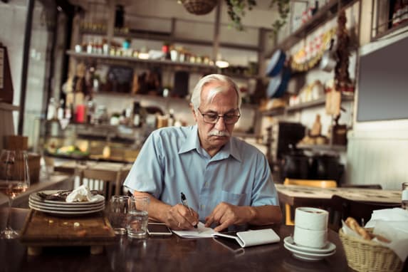 A Small Business Owner Sitting At A Table At His Restaurant, Cluttered With Dirty Dishes, Using A Calculator And Writing Stuff Down On A Little Notepad