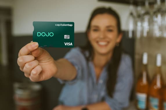 Small Business Owner In Wine Shop Holds Up The Dojo Capital On Tap Business Credit Card Smiling