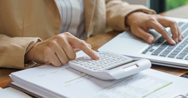 A Person Wearing A Brown Suit Jacket Sitting At A Desk With Their Laptop And A Calculator Laid On Top Of A Pile Of Invoices