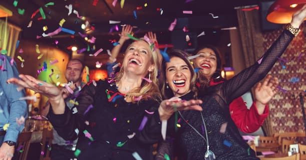 A Small Group Of Female Colleagues Smiling Into The Camera As They Get Showered With Confetting In A Dimly Lit Oub, With People Dancing In The Background