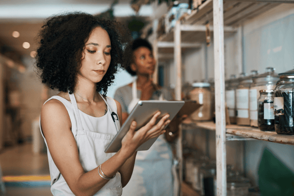 Small Business Owner Wearing An Apron Checking Her Tablet Standing Near Shelves Full Of Jars