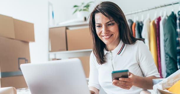 Woman Sitting At Desk With Clothes Rails Behind And Some Boxes Next To Her On The Desk, A Phone In Her Left Hand, Smiling At Her Laptop
