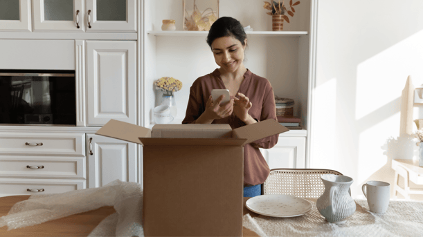 Woman Smiling Standing In Front Of Open Parcel Using Phone To Leave A Review