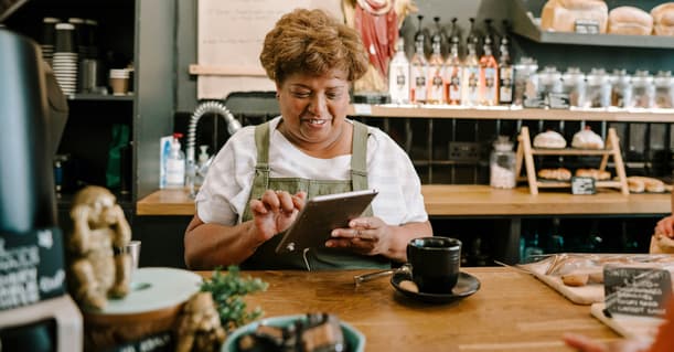A Small Business Owner In An Apron Stands Behind The Bar In A Cafe Smiling Down At Her Tablet