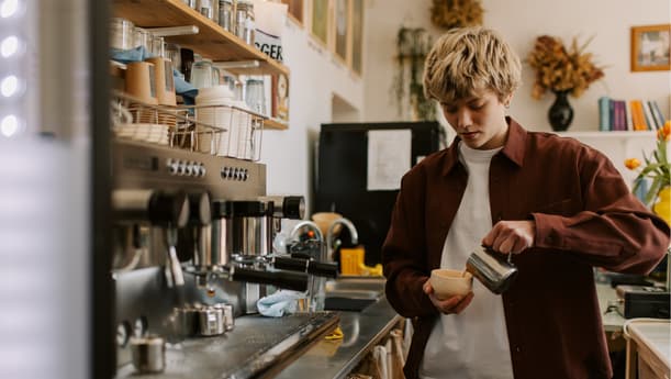 A Barista Wearing A Brown Shirt, Pouring Milk Into A Cappuccino Mug, Standing Beside A Coffee Machine In A Cafe