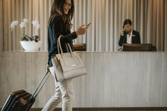 Small Business Owner With Suitcase Walks Through Hotel Lobby Looking At Mobile Phone And Smiling
