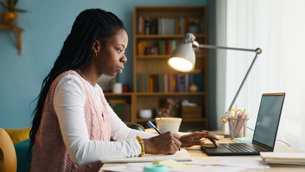 Woman Working At A Desk At Home, Writing Notes And Typing On A Laptop Under A Desk Lamp
