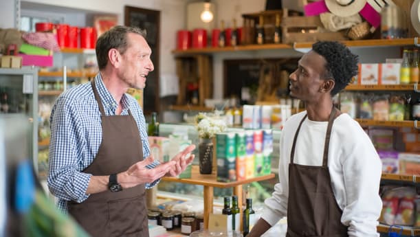Two Men In Brown Aprons Talking In A Shop Filled With Shelves Lined With Colourful Food And Produce