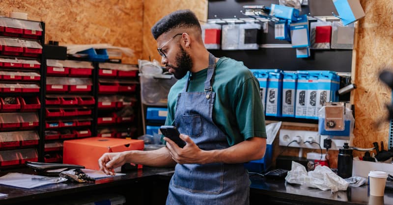 A Man In A Denim Apron Standing In Front Of Some Cluttered Shelves In A Repair Shop