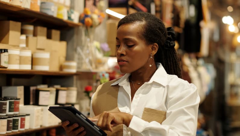 A Small Business Owner Standing In Fron Of Some Candles In A Shop Looking At Her Tablet