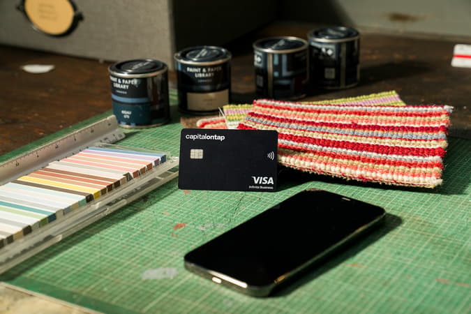 Capital On Tap Visa Card On A Designer's Cutting Mat, Surrounded By Paint Tins, Colour Swatches, Fabric Samples And A Phone