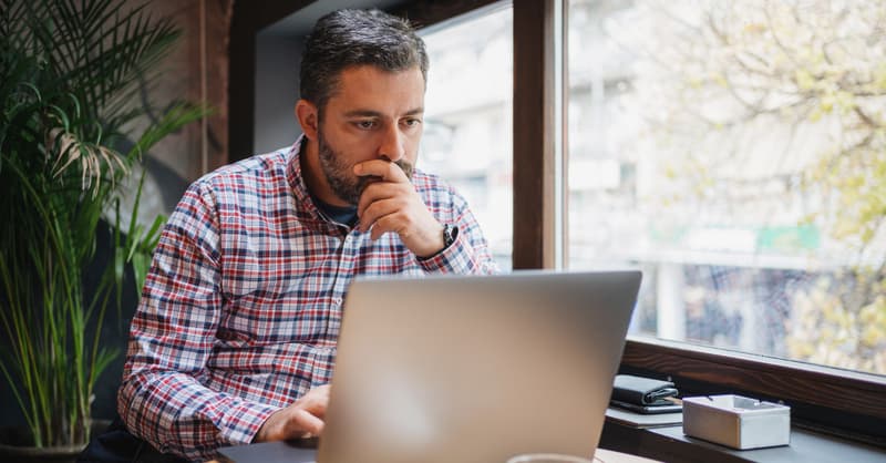 A Man In A Plaid Shirt Sitting In Front Of His Laptop At A Table Near A Window