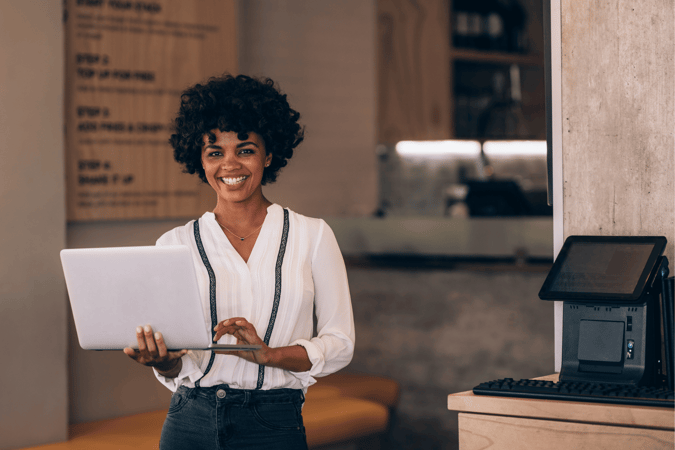 Small Business Owner Standing In A Cafe Near The Till Holding A Laptop And Smiling