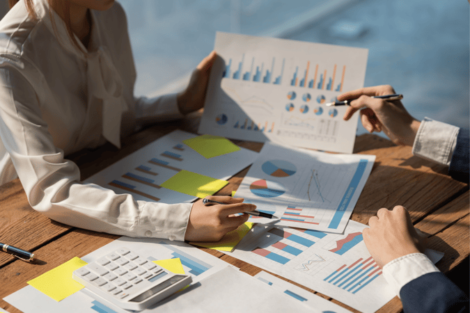 Two Employees Sitting At A Table Covered With Papers Showing Charts And Data, Trying To Figure Out Profit