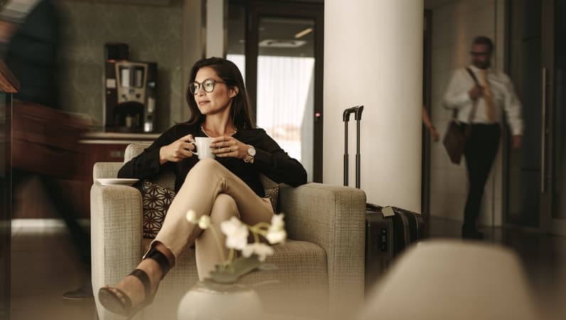 A Business Woman Sitting In An Armchair In An Airport Lounge, With Her Suitcase Nearby, Sipping From A Mug, As People Move Around In The Background
