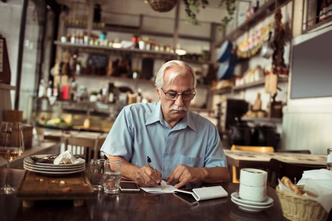 A Small Business Owner Sitting At A Table At His Restaurant, Cluttered With Dirty Dishes, Using A Calculator And Writing Stuff Down On A Little Notepad