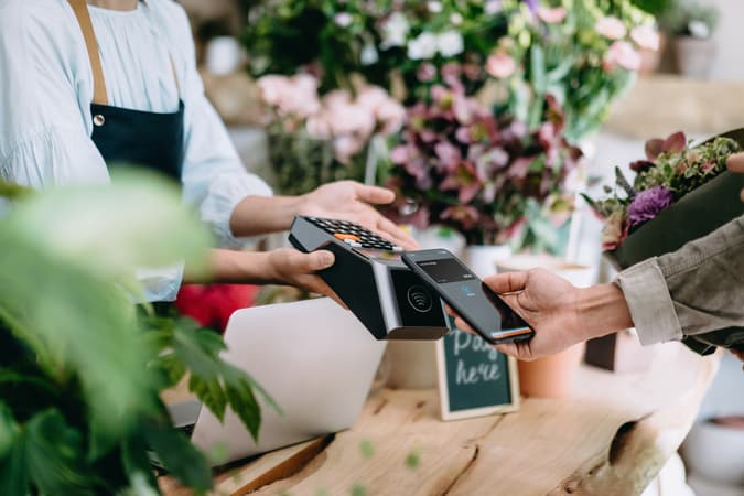 A Close Up Of A Hand Using Apple Pay With The Capital On Tap Business Credit Card At A Terminal In A Flower Shop.