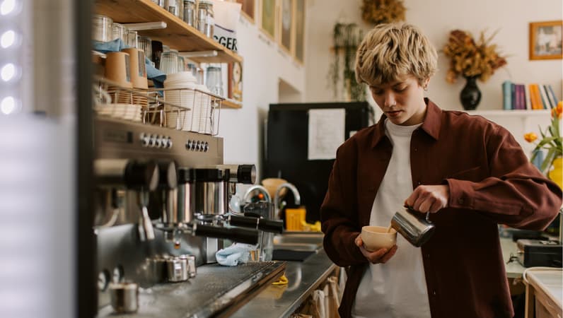 A Barista Wearing A Brown Shirt, Pouring Milk Into A Cappuccino Mug, Standing Beside A Coffee Machine In A Cafe