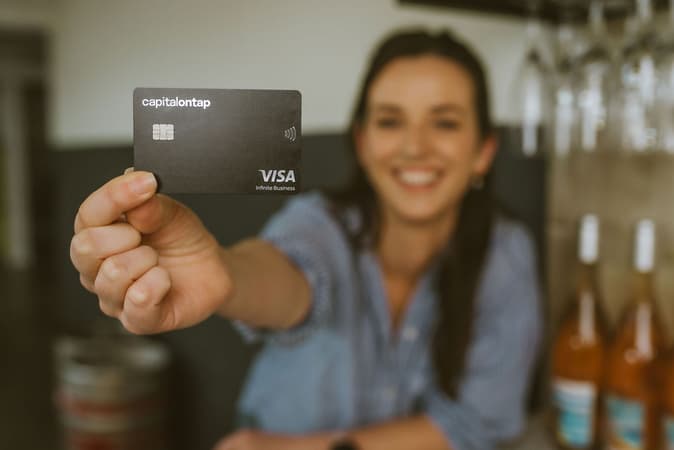 A Woman Smiling In A Wine Shop, Holding Up The Capital On Tap Business Credit Card
