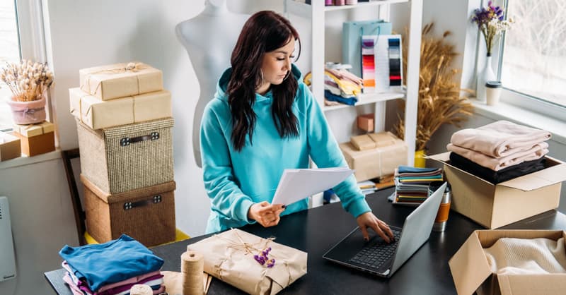 A Young Woman Standing In A Room Filled With Fabrics And Mannqeuins, At A Desk Filled With Boxes, Looking At Her Laptop,