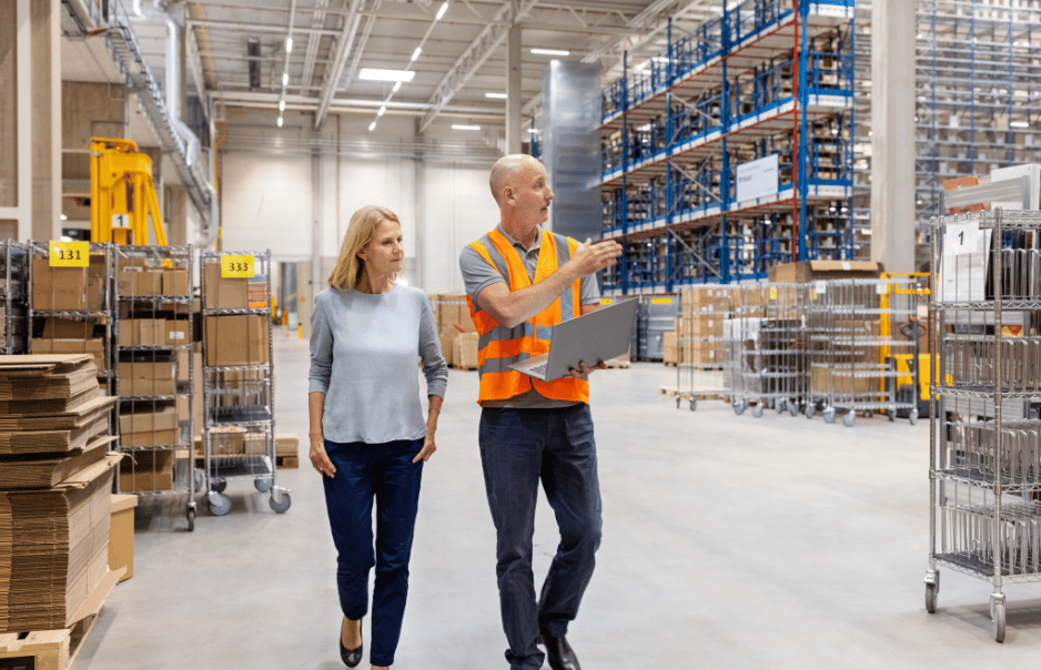 A Woman And Man In A Hi Vis Jacket Walk Through A Warehouse With Floor To Ceiling Shelves Of Stock