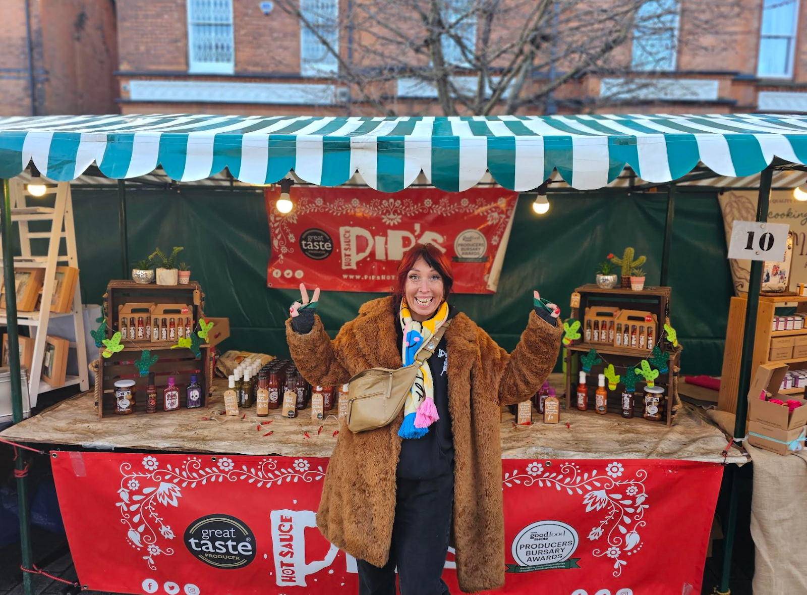 Pip's Hot Sauce owner standing in front of their Christmas Market stall, smiling, doing peace signs with their fingers