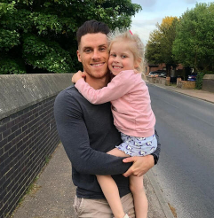 Tom is wearing a blue top and chinos and holds his young daughter, wearing a pink top and blue skirt.