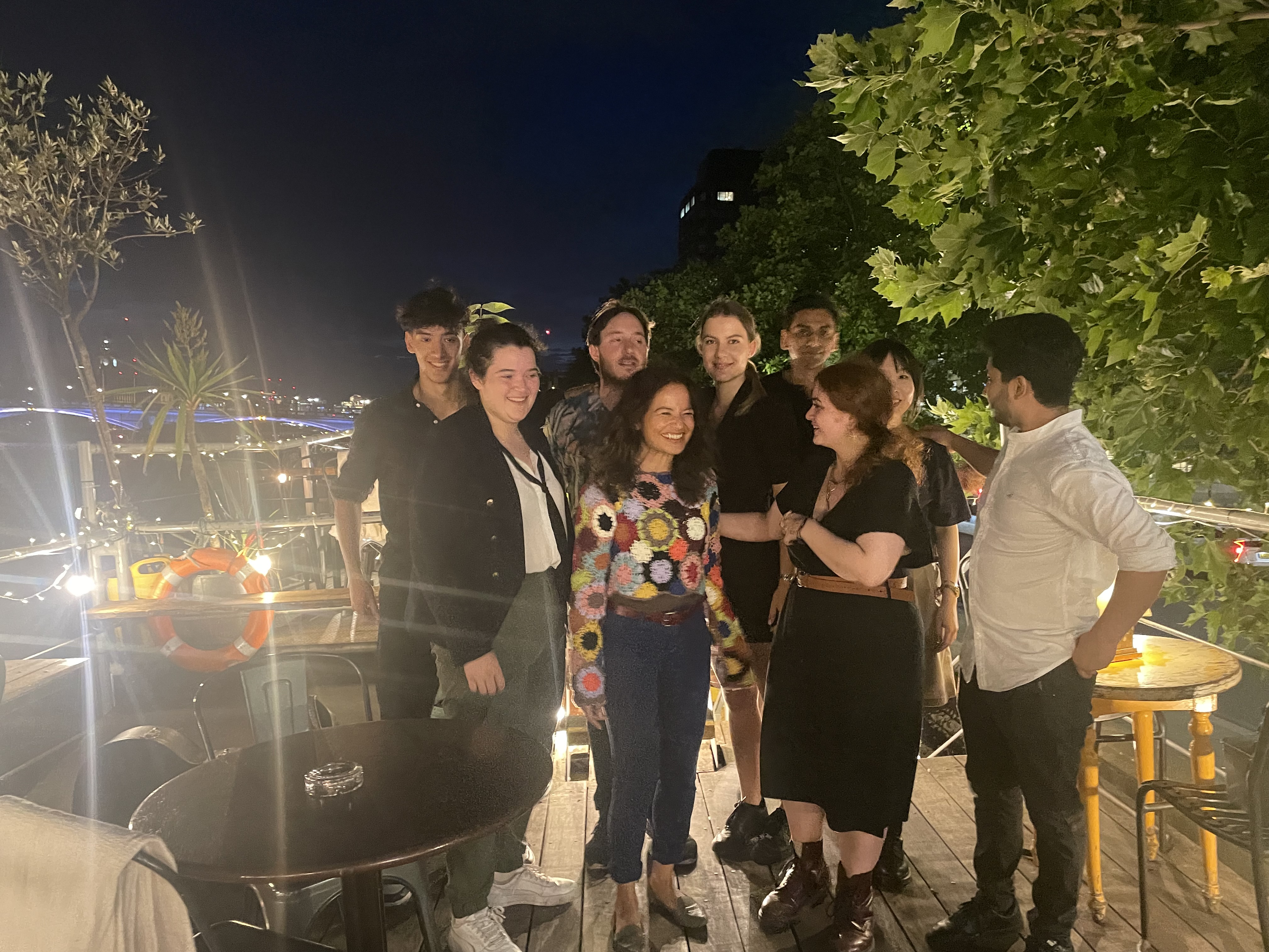 A group of friends standing together on top of the barge in the thames. It is night time, they are lit up by fairy lights