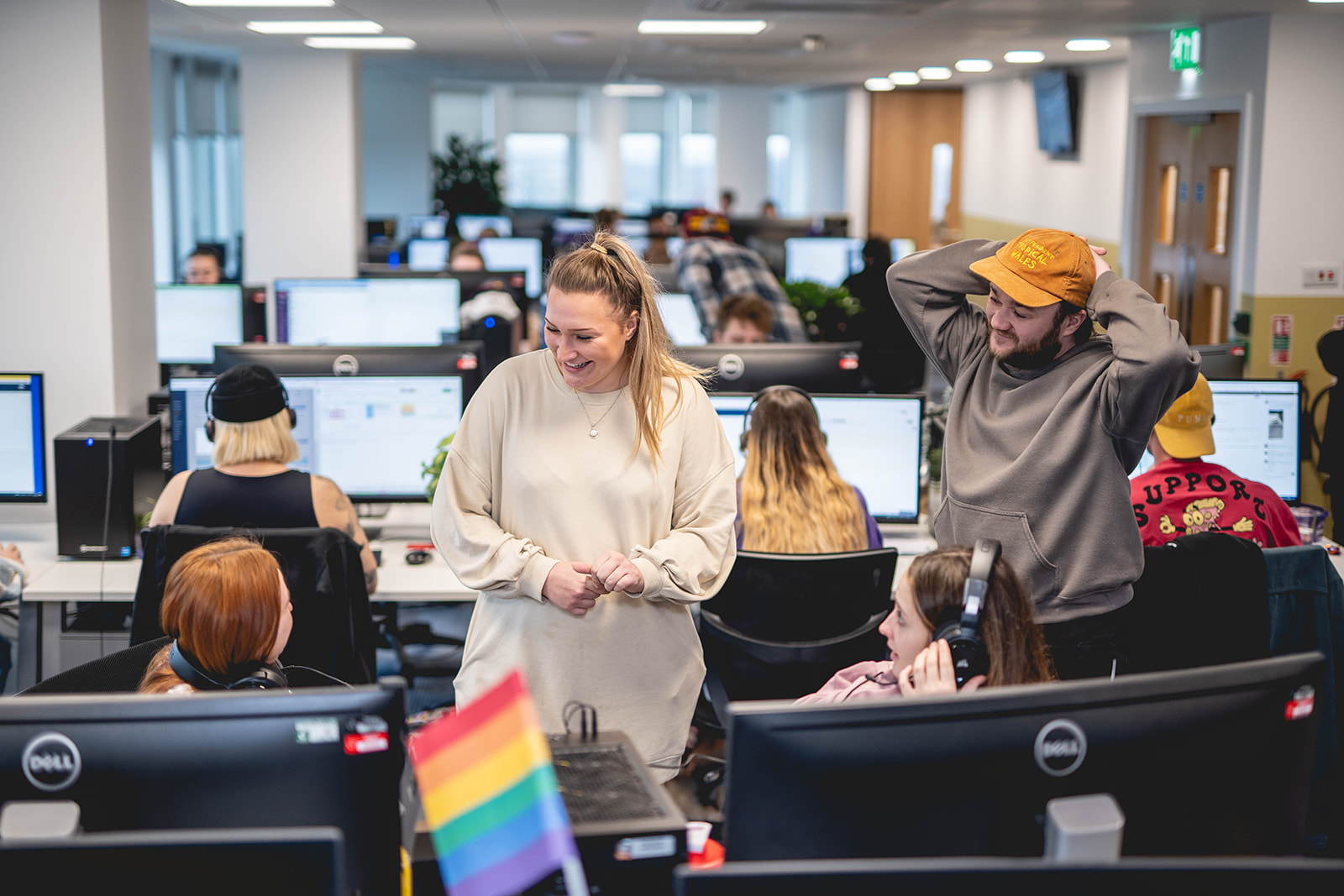 An office floor full of people working on computers. 4 people are talking at the forefront